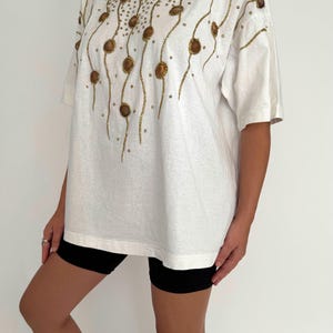 May include: White oversized t-shirt with a gold and brown seashell design. The design features a repeating pattern of seashells and gold glitter.