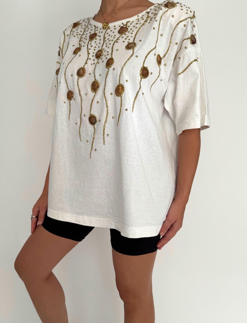 May include: White oversized t-shirt with a gold and brown seashell design. The design features a repeating pattern of seashells and gold glitter.