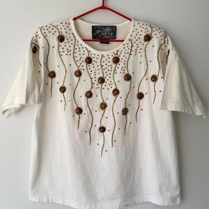 May include: White short-sleeved t-shirt with a gold and brown beaded design. The design features a series of wavy lines with small brown circles and gold beads. The t-shirt has the text "Apparel Unlimited" and "Show Wear" on a black tag.