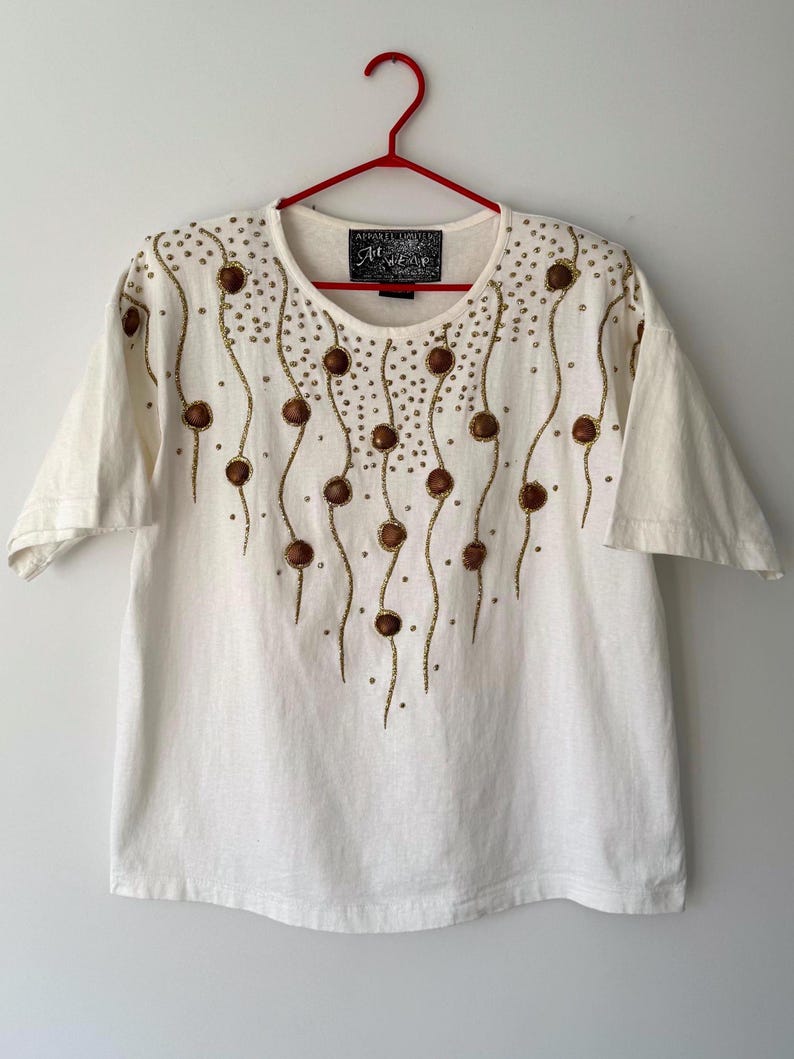 May include: White short-sleeved t-shirt with a gold and brown beaded design. The design features a series of wavy lines with small brown circles and gold beads. The t-shirt has the text "Apparel Unlimited" and "Show Wear" on a black tag.