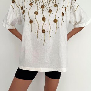 May include: White oversized t-shirt with a gold and brown beaded design. The design features a series of curved lines with small gold beads and larger brown beads.