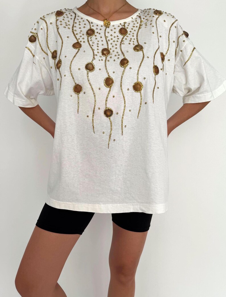 May include: White oversized t-shirt with a gold and brown beaded design. The design features a series of curved lines with small gold beads and larger brown beads.