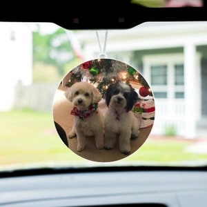 May include: A round car air freshener with a photo of two white dogs in front of a Christmas tree. The dogs are wearing red and white collars.