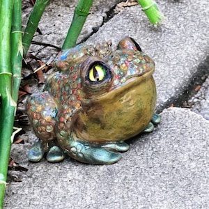 Puede incluir: Una figurita de rana de cerámica pintada en verde, marrón y dorado. La rana tiene ojos grandes y amarillos y una superficie texturizada.