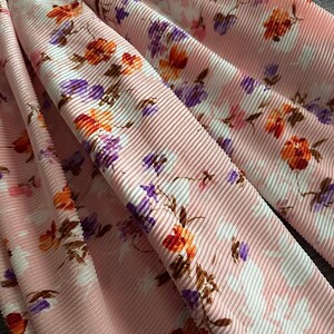 May include: A light pink pleated fabric featuring a floral pattern with orange, purple, and white flowers. The material has a distinct vertical ribbed texture throughout, creating deep folds and shadows across the surface of the textile.