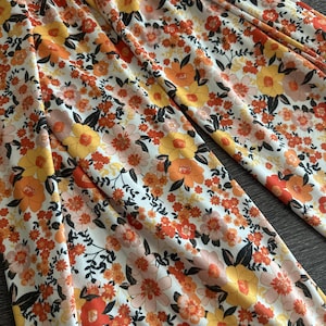 May include: Close-up of a fabric with a floral pattern. The design features a white background with orange, yellow, and peach-colored flowers, accented by black leaves. The fabric appears to be lightweight and suitable for clothing.