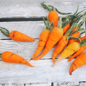 Könnte beinhalten: Eine Sammlung handgefertigter orangefarbener Filzkarotten mit grünen Stielen, angeordnet auf einer verwitterten, weißen Holzoberfläche. Die Karotten variieren leicht in Größe und Form und zeigen eine rustikale, natürliche Ästhetik. Die Filzstruktur verleiht der Gemüse-Dekoration eine weiche, taktile Qualität.