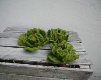 Può includere: Tre sculture fatte a mano in feltro verde a forma di cavolo con accenti gialli poggiano su una superficie di legno grigio e invecchiato. I pezzi in feltro hanno un aspetto strutturato e organico, che ricorda cavoli in miniatura o elementi floreali decorativi.