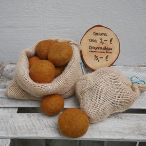 Könnte beinhalten: Zwei Jutesäcke gefüllt mit Filzkugeln, ein Sack ist offen und die Kugeln sind sichtbar. Die Kugeln sind tief orangefarben. Ein Holzschild mit dem Text "Kartoffel Stick 2,- € Kartoffelsticks 15,- €" ist im Hintergrund sichtbar.