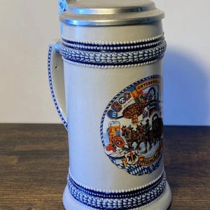 May include: A white ceramic beer stein with a metal lid and handle. The stein features blue trim and an oval graphic with the word "Oktoberfest" and a scene of horses and people. The stein is on a wooden surface.