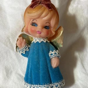 May include: Vintage angel Christmas ornament. The angel has a light skin tone, blonde hair, and blue eyes. She wears a blue dress with white lace trim and gold wings. A brown bow adorns her head.