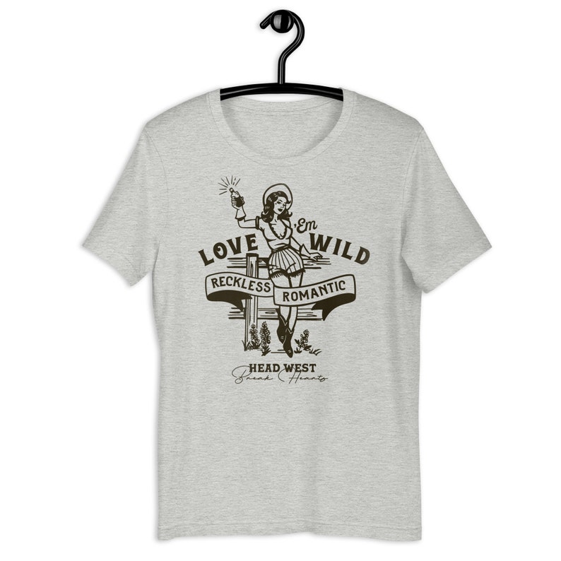 May include: A grey t-shirt with a vintage-style illustration of a woman in a cowboy hat holding a bottle. The text "Love 'Em Wild" is above the woman, with the words "Reckless" and "Romantic" on banners below her. The text "Head West Break Hearts" is at the bottom of the illustration.