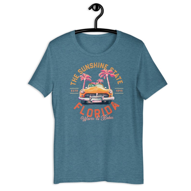 Puede incluir: Una camiseta azul turquesa con un gr&aacute;fico de estilo vintage de un coche amarillo conduciendo a trav&eacute;s de palmeras. El gr&aacute;fico dice "The Sunshine State, Est. 1845, Florida, Waves & Babes".
