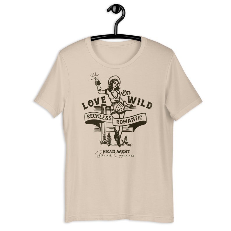 May include: A beige t-shirt with a graphic print of a woman in a cowboy hat holding a bottle. The graphic says "Love 'Em Wild" with the words "Reckless" and "Romantic" on a banner below. The text "Head West" and "Break Hearts" is below the banner.