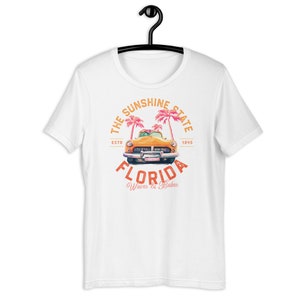 Puede incluir: Camiseta blanca con un gr&aacute;fico de estilo vintage de un coche amarillo conduciendo a trav&eacute;s de palmeras. El gr&aacute;fico dice "The Sunshine State, ESTD 1845, FLORIDA, Waves & Babes".