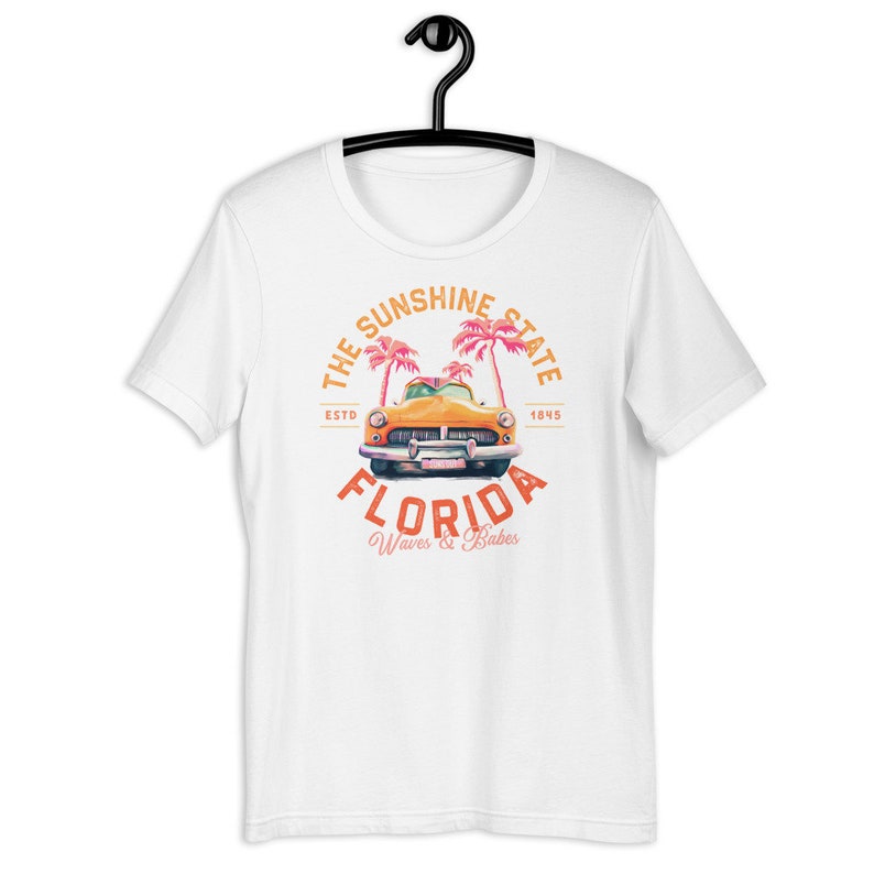Puede incluir: Camiseta blanca con un gr&aacute;fico de estilo vintage de un coche amarillo conduciendo a trav&eacute;s de palmeras. El gr&aacute;fico dice "The Sunshine State, ESTD 1845, FLORIDA, Waves & Babes".