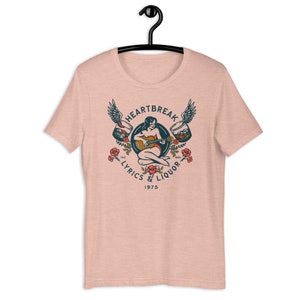 May include: A pink t-shirt with a vintage-style graphic design featuring a woman playing a guitar with wings and glasses of liquor on either side. The text reads "Heartbreak Lyrics & Liquor 1975".