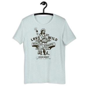 May include: Light blue t-shirt with a vintage-style illustration of a woman in a cowboy hat holding a bottle. The text "Love 'Em Wild" is above the illustration, with the words "Reckless" and "Romantic" on banners below. The text "Head West Break Hearts" is at the bottom of the design.