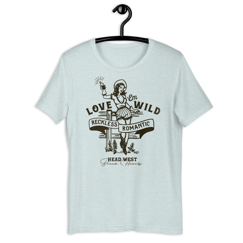 May include: Light blue t-shirt with a vintage-style illustration of a woman in a cowboy hat holding a bottle. The text "Love 'Em Wild" is above the illustration, with the words "Reckless" and "Romantic" on banners below. The text "Head West Break Hearts" is at the bottom of the design.