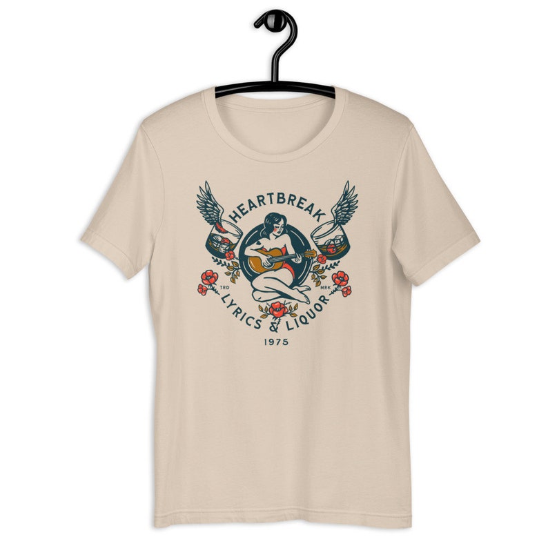 May include: A beige t-shirt with a vintage-style graphic design featuring a woman playing a guitar with wings and glasses of liquor. The text reads "Heartbreak Lyrics & Liquor 1975".