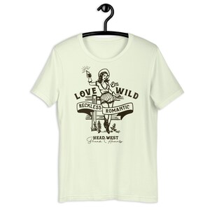May include: A light green t-shirt with a vintage-style illustration of a woman in a cowboy hat holding a bottle. The text "Love 'Em Wild" is above the illustration, with the words "Reckless" and "Romantic" on banners below. The text "Head West Break Hearts" is at the bottom of the design.