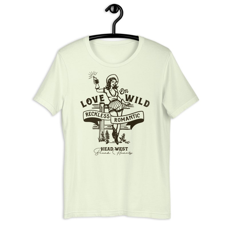May include: A light green t-shirt with a vintage-style illustration of a woman in a cowboy hat holding a bottle. The text "Love 'Em Wild" is above the illustration, with the words "Reckless" and "Romantic" on banners below. The text "Head West Break Hearts" is at the bottom of the design.