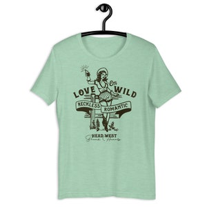 May include: A light green t-shirt with a vintage-style illustration of a woman holding a bottle and a gun. The text "Love 'Em Wild" is above the woman, and the text "Reckless Romantic" is below her. The text "Head West" is at the bottom of the illustration.
