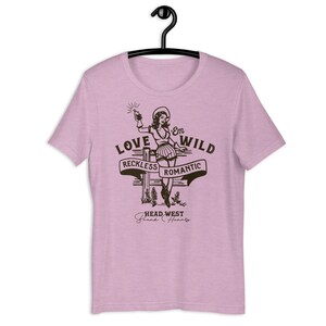 May include: A pink t-shirt with a graphic of a woman in a cowboy hat holding a bottle of liquor. The graphic says "Love 'Em Wild, Reckless Romantic, Head West, Break Hearts".