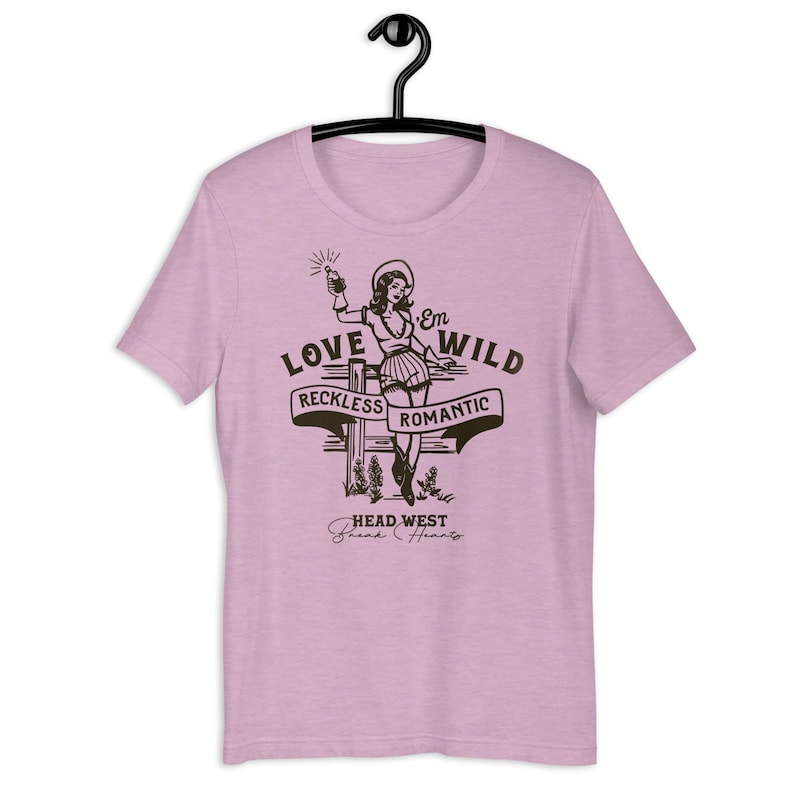 May include: A pink t-shirt with a graphic of a woman in a cowboy hat holding a bottle of liquor. The graphic says "Love 'Em Wild, Reckless Romantic, Head West, Break Hearts".