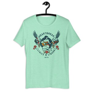 May include: A light green t-shirt with a vintage-style graphic design featuring a woman playing a guitar with wings and glasses of liquor. The text reads "Heartbreak Lyrics & Liquor 1975".
