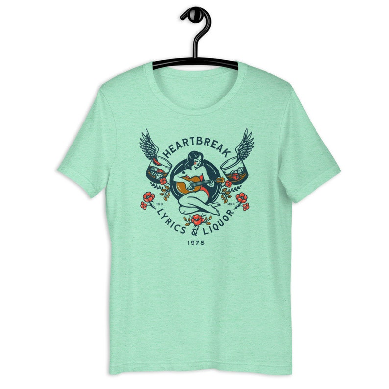 May include: A light green t-shirt with a vintage-style graphic design featuring a woman playing a guitar with wings and glasses of liquor. The text reads "Heartbreak Lyrics & Liquor 1975".