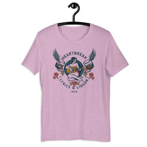 May include: Pink t-shirt with a vintage-style graphic of a woman playing a guitar with wings and glasses of liquor on either side. The text reads "Heartbreak Lyrics & Liquor 1975".