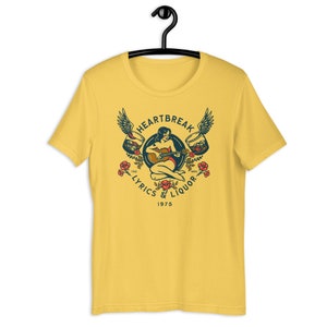 May include: Yellow t-shirt with a vintage-style illustration of a woman playing a guitar with wings and glasses of liquor on either side. The text "Heartbreak Lyrics & Liquor 1975" is printed below the illustration.