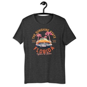 Puede incluir: Una camiseta gris oscuro jaspeada con un gr&aacute;fico de estilo vintage de un coche amarillo conduciendo a trav&eacute;s de palmeras. El gr&aacute;fico incluye el texto "The Sunshine State, Est. 1845, Florida, Waves & Babes".