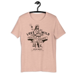 May include: A pink t-shirt with a graphic of a woman in a cowboy hat holding a bottle and a gun. The graphic says "Love 'Em Wild" with the words "Reckless" and "Romantic" on a banner below. The graphic also says "Head West" with the words "Break Hearts" below.