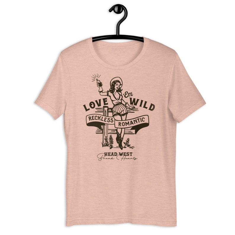 May include: A pink t-shirt with a graphic of a woman in a cowboy hat holding a bottle and a gun. The graphic says "Love 'Em Wild" with the words "Reckless" and "Romantic" on a banner below. The graphic also says "Head West" with the words "Break Hearts" below.
