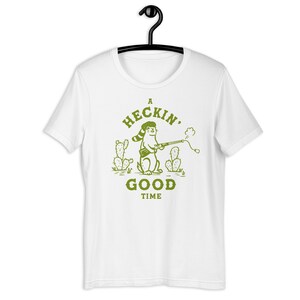 Könnte beinhalten: Weißes T-Shirt mit einer grünen Illustration eines Comic-Tieres, das einen Hut trägt und ein Gewehr hält. Der Text "A Heck'n Good Time" ist unter der Illustration gedruckt.