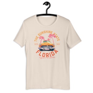 Puede incluir: Una camiseta beige con un dise&ntilde;o gr&aacute;fico de estilo vintage que presenta un coche amarillo conduciendo a trav&eacute;s de palmeras. El texto dice "The Sunshine State, Est. 1845, Florida, Waves & Babes".