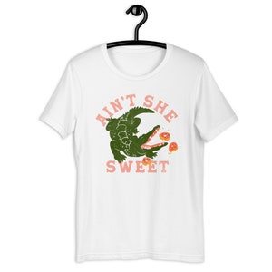 May include: White t-shirt with a green alligator eating pink donuts and the text "Ain't She Sweet" in pink.