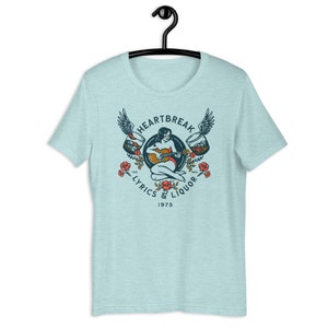 May include: A light blue t-shirt with a vintage-style graphic design featuring a woman playing a guitar with wings and two glasses of liquor. The text reads "Heartbreak Lyrics & Liquor 1975".