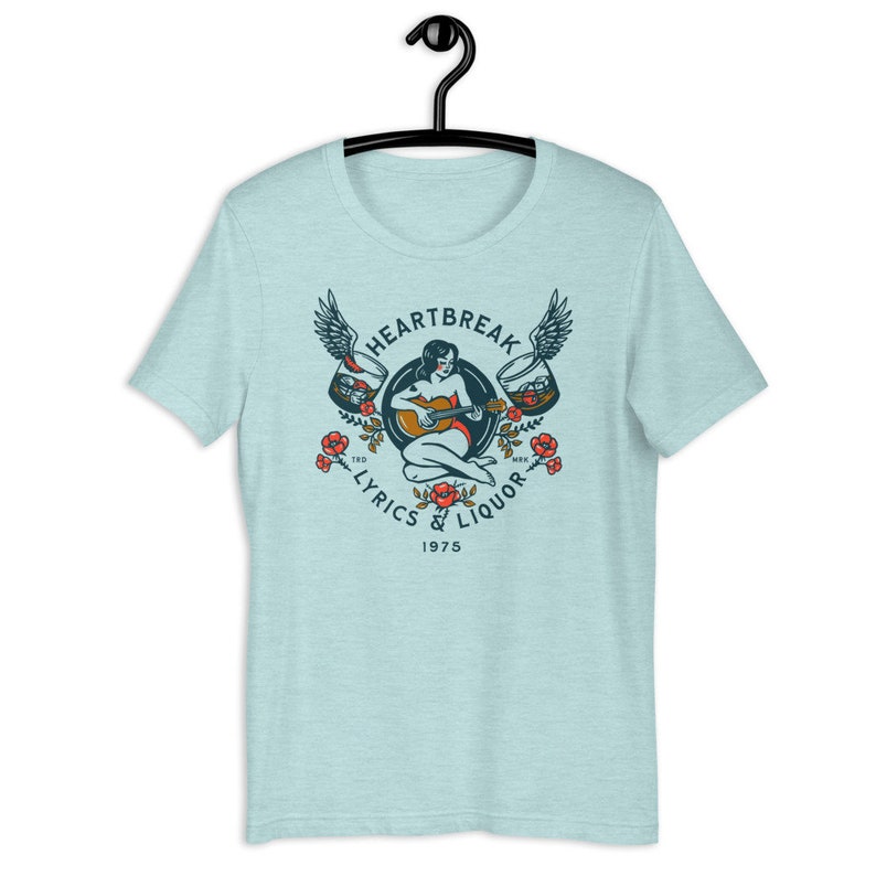 May include: A light blue t-shirt with a vintage-style graphic design featuring a woman playing a guitar with wings and two glasses of liquor. The text reads "Heartbreak Lyrics & Liquor 1975".