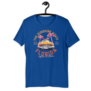 Puede incluir: Una camiseta azul real con un gr&aacute;fico de estilo vintage de un coche amarillo conduciendo a trav&eacute;s de palmeras. El gr&aacute;fico dice "The Sunshine State, Florida, Est. 1845, Waves & Babes".