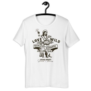 May include: White t-shirt with a black and white illustration of a woman holding a bottle. The text "Love 'Em Wild" is above the woman, with the words "Reckless" and "Romantic" on banners below her. The text "Head West Break Hearts" is below the woman.
