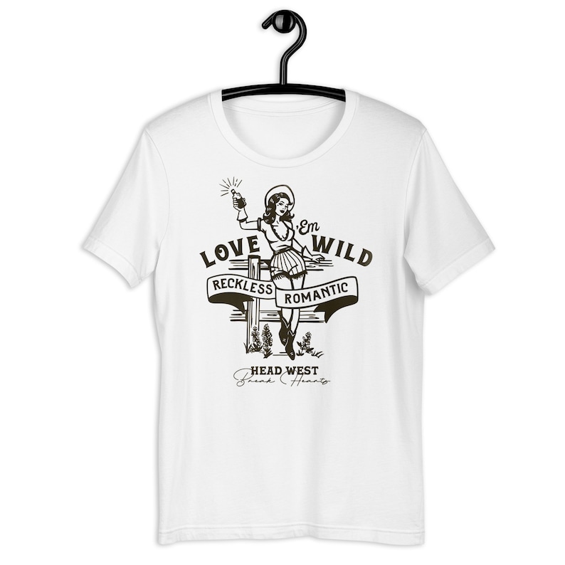 May include: White t-shirt with a black and white illustration of a woman holding a bottle. The text "Love 'Em Wild" is above the woman, with the words "Reckless" and "Romantic" on banners below her. The text "Head West Break Hearts" is below the woman.