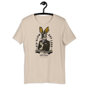 Puede incluir: Una camiseta beige con un gráfico de un conejo con traje y sosteniendo un vaso de bourbon. El texto en la camiseta dice "Sip Slow Say Less Mister Bourbon Drink Like a Gentleman Whiskey Reign".