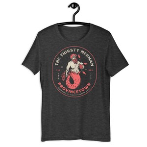 Peut inclure: Un t-shirt gris foncé chiné avec un graphique circulaire d'un homme-poisson rouge tenant une chope de bière. Le graphique indique "The Thirsty Merman" et "Provincetown Purveyors of Finely Crafted Ales".
