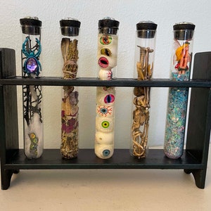 Może przedstawiać: Sześć szklanych probówek wypełnionych różnymi przedmiotami o tematyce Halloween, w tym pająkiem, oczami, brokatem i wróżką. Probówki są ułożone w czarnym drewnianym stojaku.