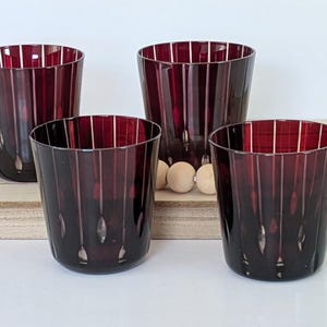 May include: A set of four deep burgundy glass tumblers featuring a vertical ribbed texture. Each tumbler has a cylindrical shape with a slightly tapered base and a smooth, clear rim, showcasing a dark, translucent red color throughout the glass.
