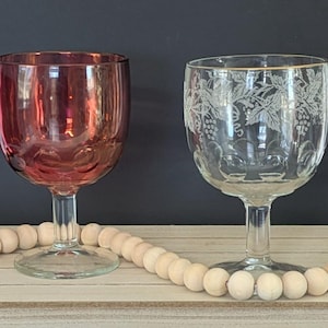 May include: Two wine glasses. One is a deep red color, and the other is clear with a gold rim and etched grape vine design. The glasses sit on a wooden surface with a strand of wooden beads in front of them.