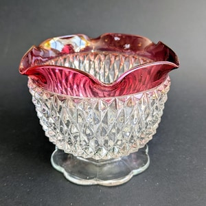 May include: A clear glass bowl with a ruby red rim and a diamond-cut pattern. The bowl sits on a clear, scalloped base. The rim has a wavy design. The bowl is likely vintage or antique.